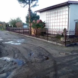 Metalowe ogrodzenie posesji z ozdobnymi elementami, widoczne z błotnistej drogi, w tle dom z rusztowaniem na ścianie.