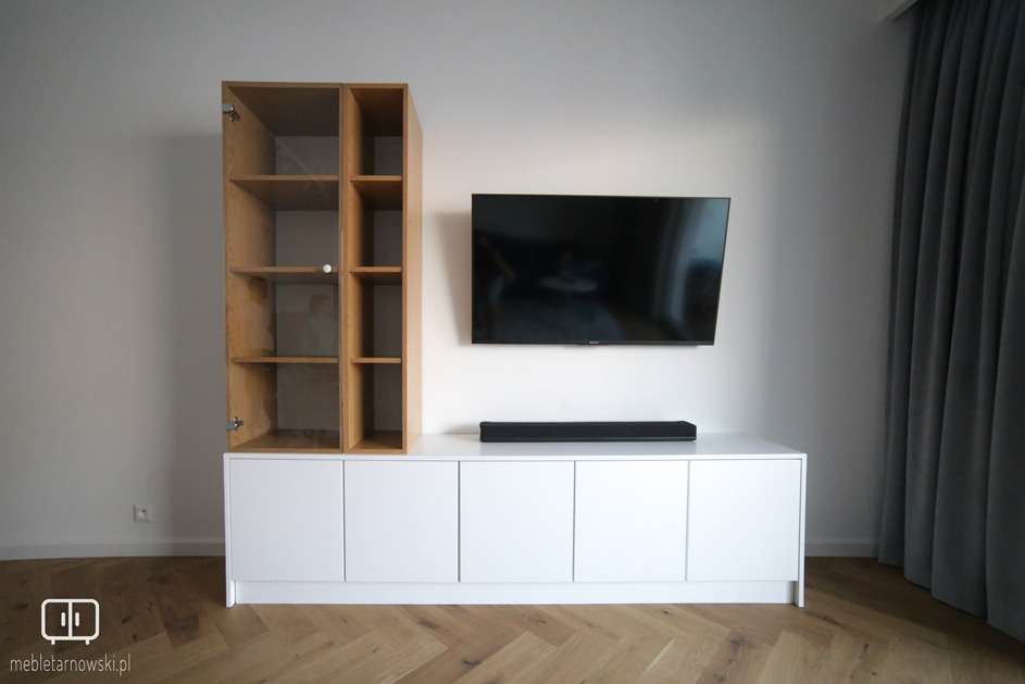 Biała szafka RTV z szafką z otwartymi półkami z jasnego drewna, wiszący telewizor i listwa głośnikowa na tle białej ściany i parkietu w jodełkę.
