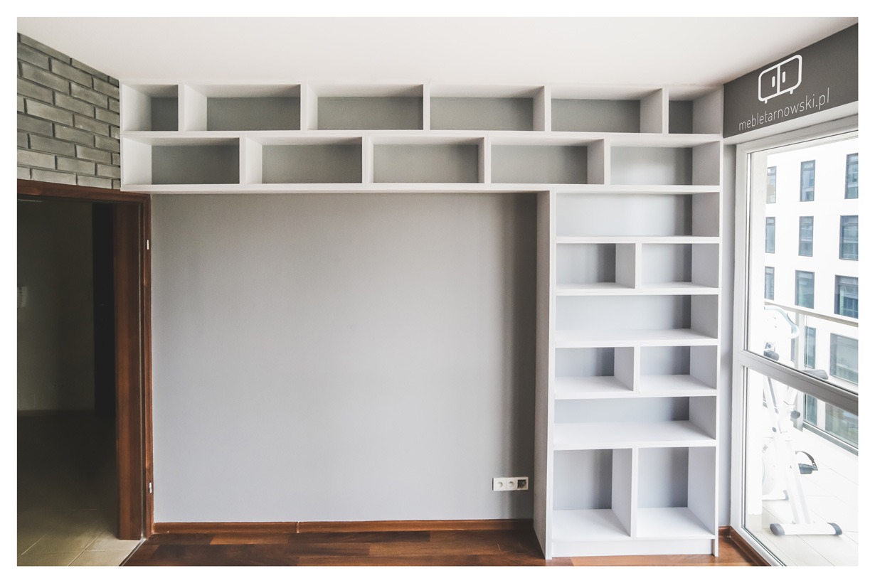 Biała zabudowa regałowa z półkami nad drzwiami, zintegrowana z szafą wnękową, widok na szarą ścianę i parkiet, logo firmy na szybie okna.