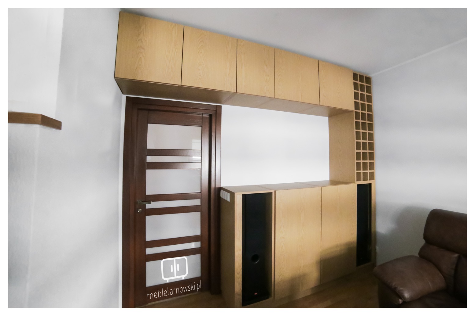 Zabudowa ściany w salonie z jasnego drewna, obejmująca szafki wiszące, regał na wino i szafki pod telewizor, zintegrowana z drzwiami wewnętrznymi.