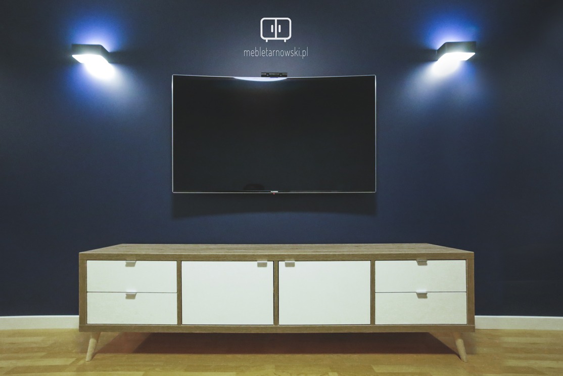 Nowoczesna szafka RTV z białymi frontami szuflad i drewnianym blatem, ustawiona pod telewizorem na ciemnoniebieskiej ścianie z dwoma kinkietami.