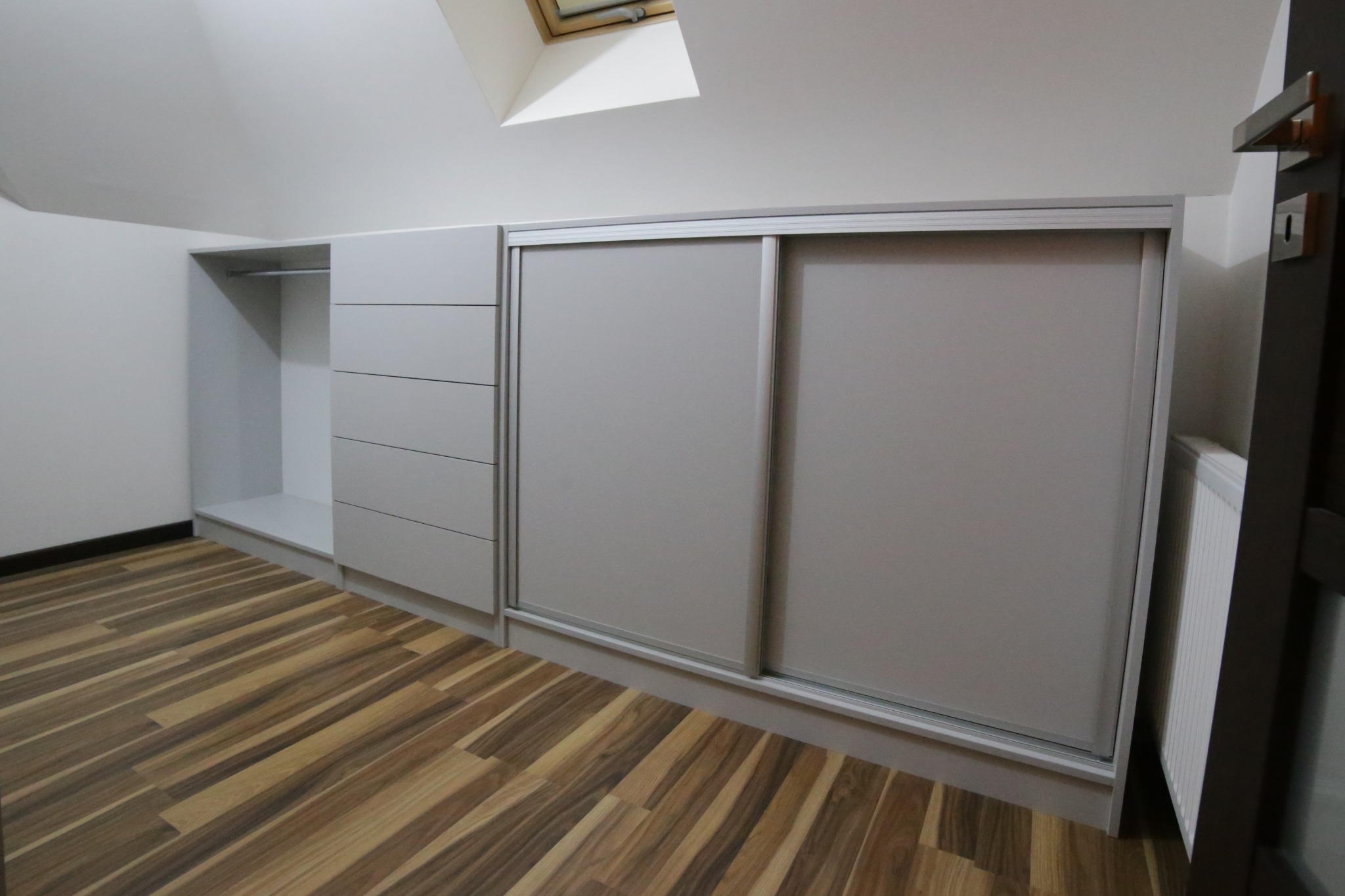 Szara zabudowa meblowa pod skosem z przesuwnymi drzwiami i szufladami, widok na drewnianą podłogę i fragment okna dachowego.