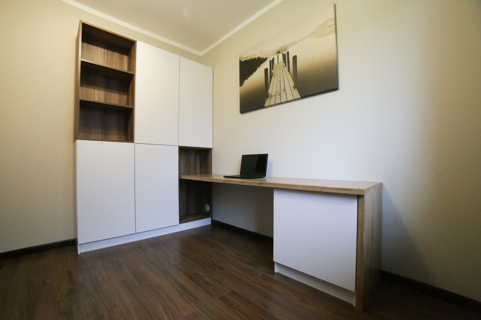 Białe szafy z drewnianymi akcentami, biurko z laptopem, obraz z molo na ścianie, brązowa podłoga.