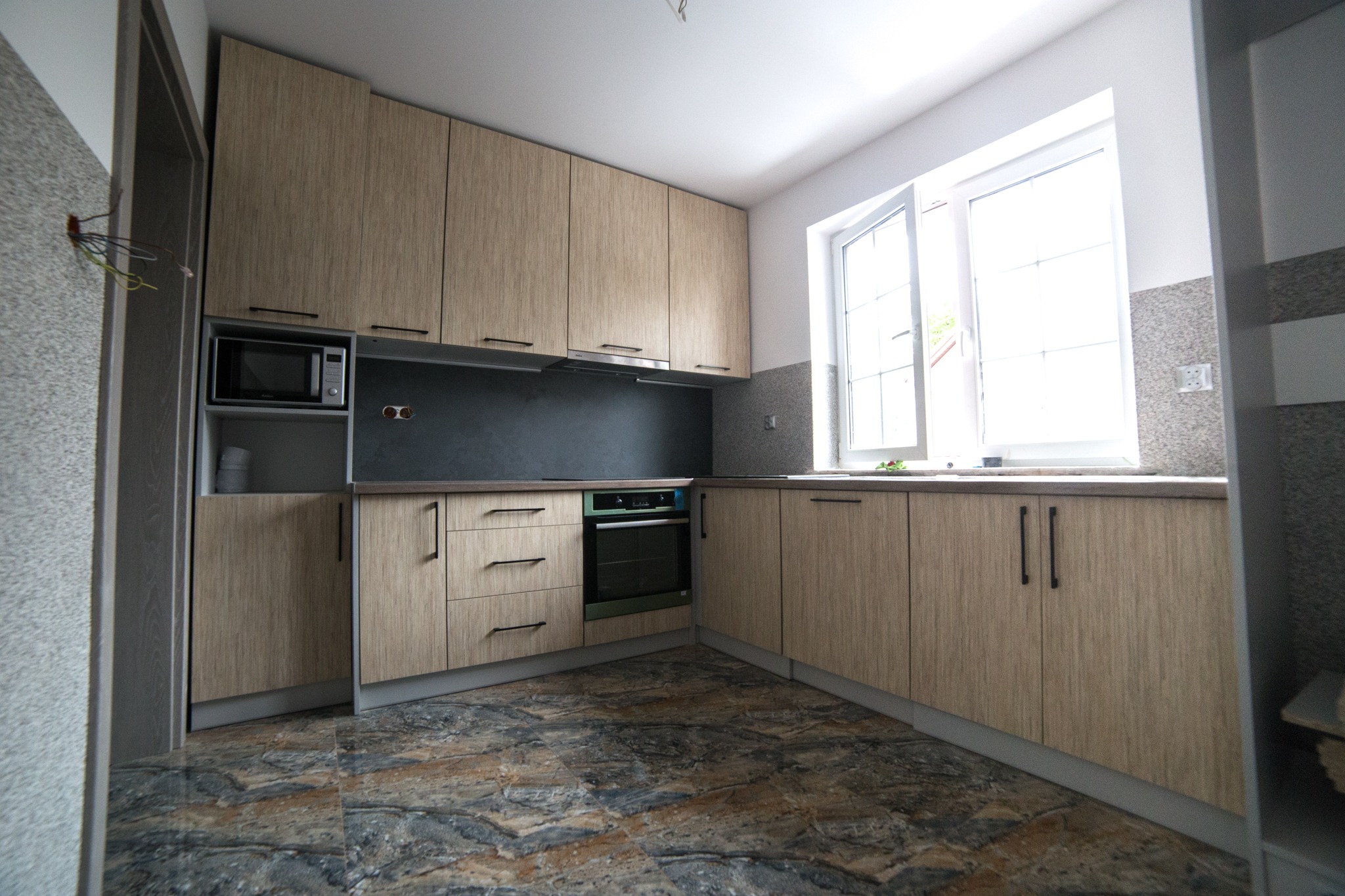 Jasna kuchnia z drewnianymi szafkami na wymiar, czarnymi uchwytami i ciemnym blatem, widoczne gniazdka elektryczne nad blatem i odsłonięte przewody po lewej stronie.