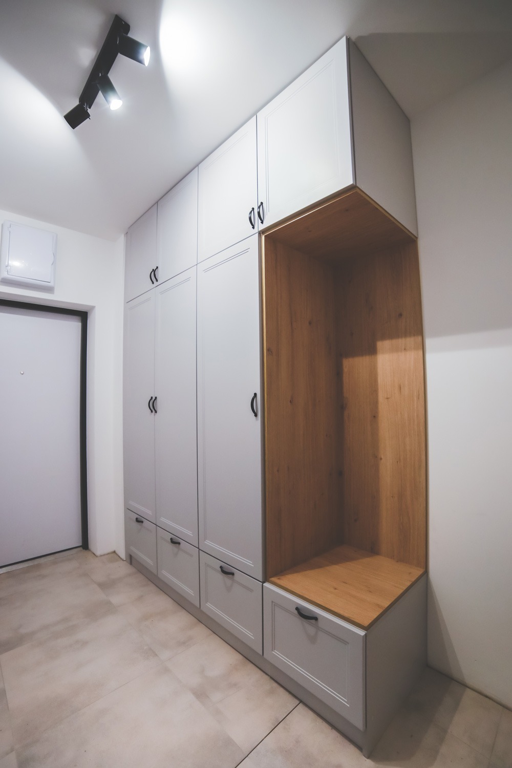 Szara zabudowa w przedpokoju z szafami i otwartą wnęką z drewnianym wykończeniem, czarne uchwyty, oświetlenie punktowe na suficie.