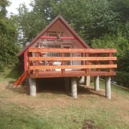 Mały, drewniany domek letniskowy z tarasem, wsparty na betonowych słupach, otoczony zielenią drzew i krzewów. Widoczny fragment zjeżdżalni.