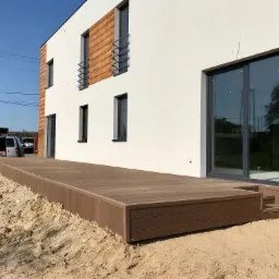 Nowoczesny taras z deski kompozytowej w kolorze brązu, przylegający do białego budynku z drewnopodobnymi elementami dekoracyjnymi, widok z poziomu gruntu na tle nieba.