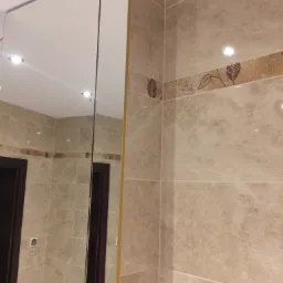 Beżowe płytki marmurowe z dekoracyjnym paskiem z motywem liści na ścianie łazienki, odbicie w lustrzanej szafce nad umywalką.