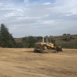 Żółty spychacz na wyrównanym terenie z widocznymi śladami pracy, drzewa w tle i pochmurne niebo.