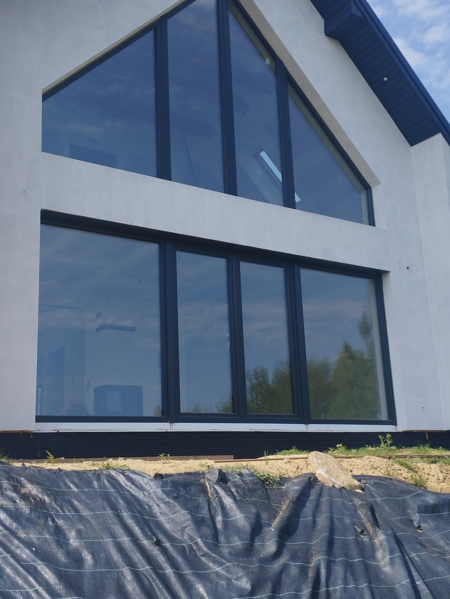 Nowoczesna elewacja domu z dużymi, prostokątnymi oknami z czarnymi ramami i trójkątnym oknem na poddaszu. Na pierwszym planie czarna folia budowlana.