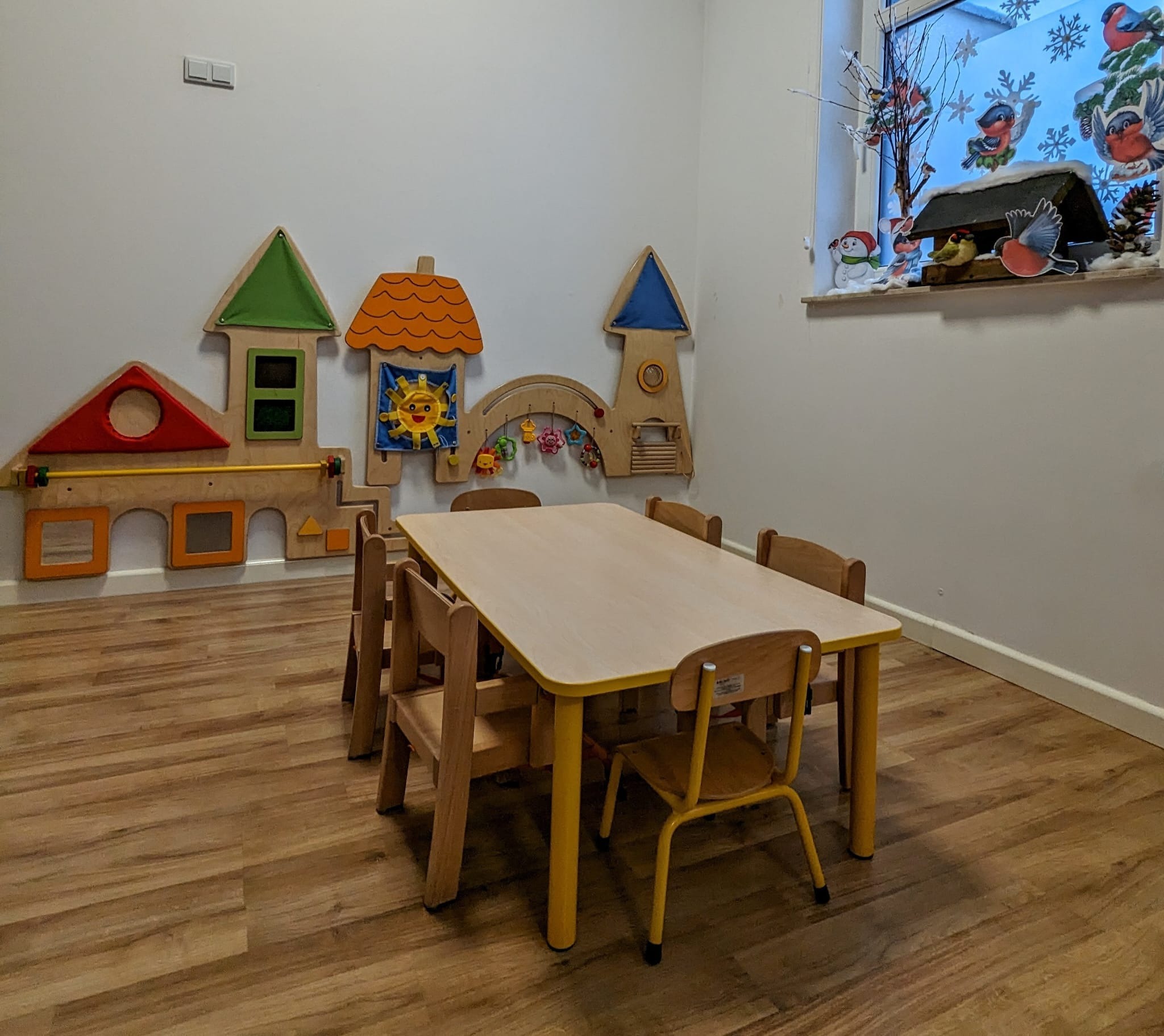 Sala zabaw z drewnianym stołem i krzesłami, dekoracjami ściennymi w kształcie domków i zimową dekoracją okienną z ptakami