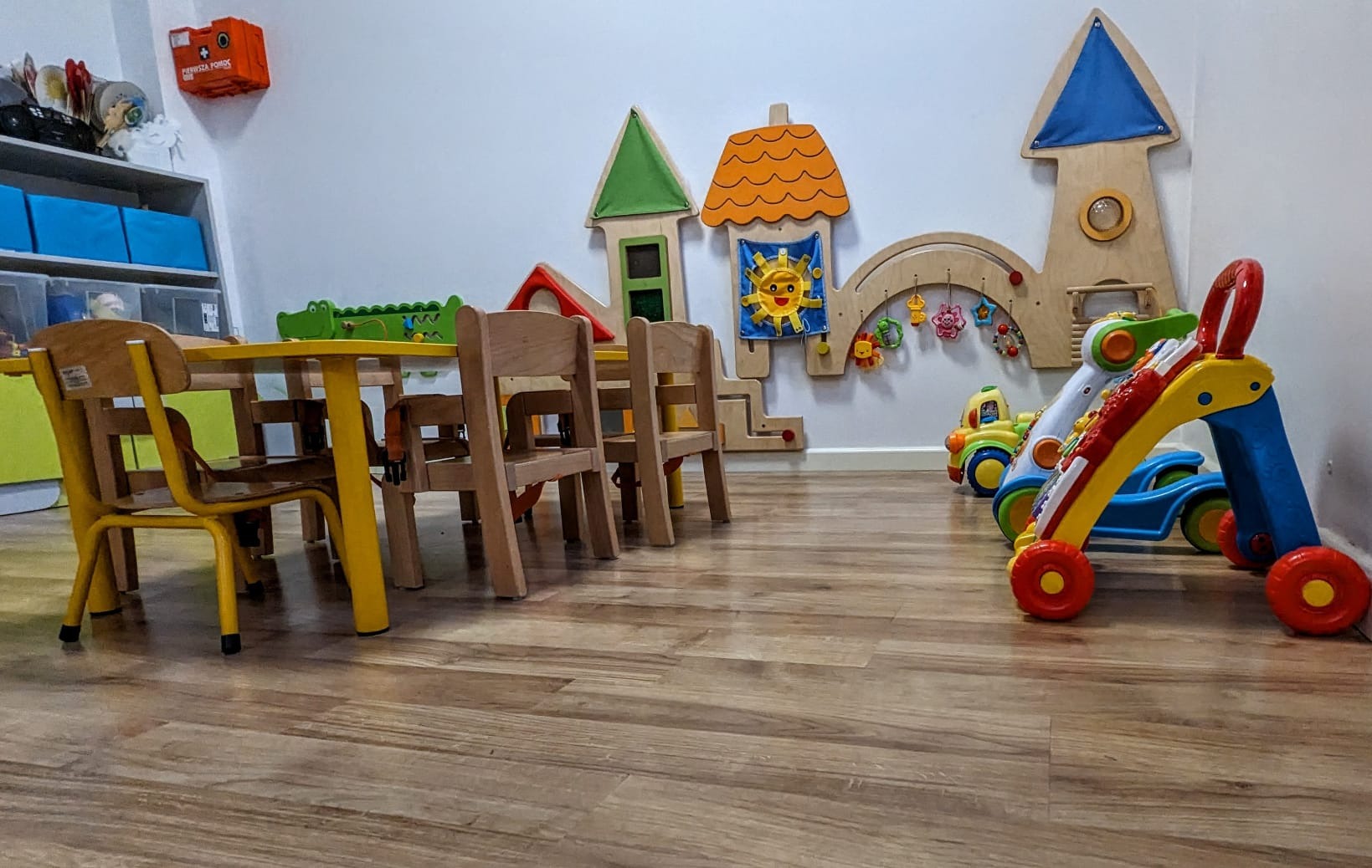 Wnętrze sali zabaw z niskimi, żółtymi stolikami i krzesłami, drewnianymi zabawkami na ścianie oraz chodzikami dla dzieci w kolorach niebieskim, żółtym i czerwonym.