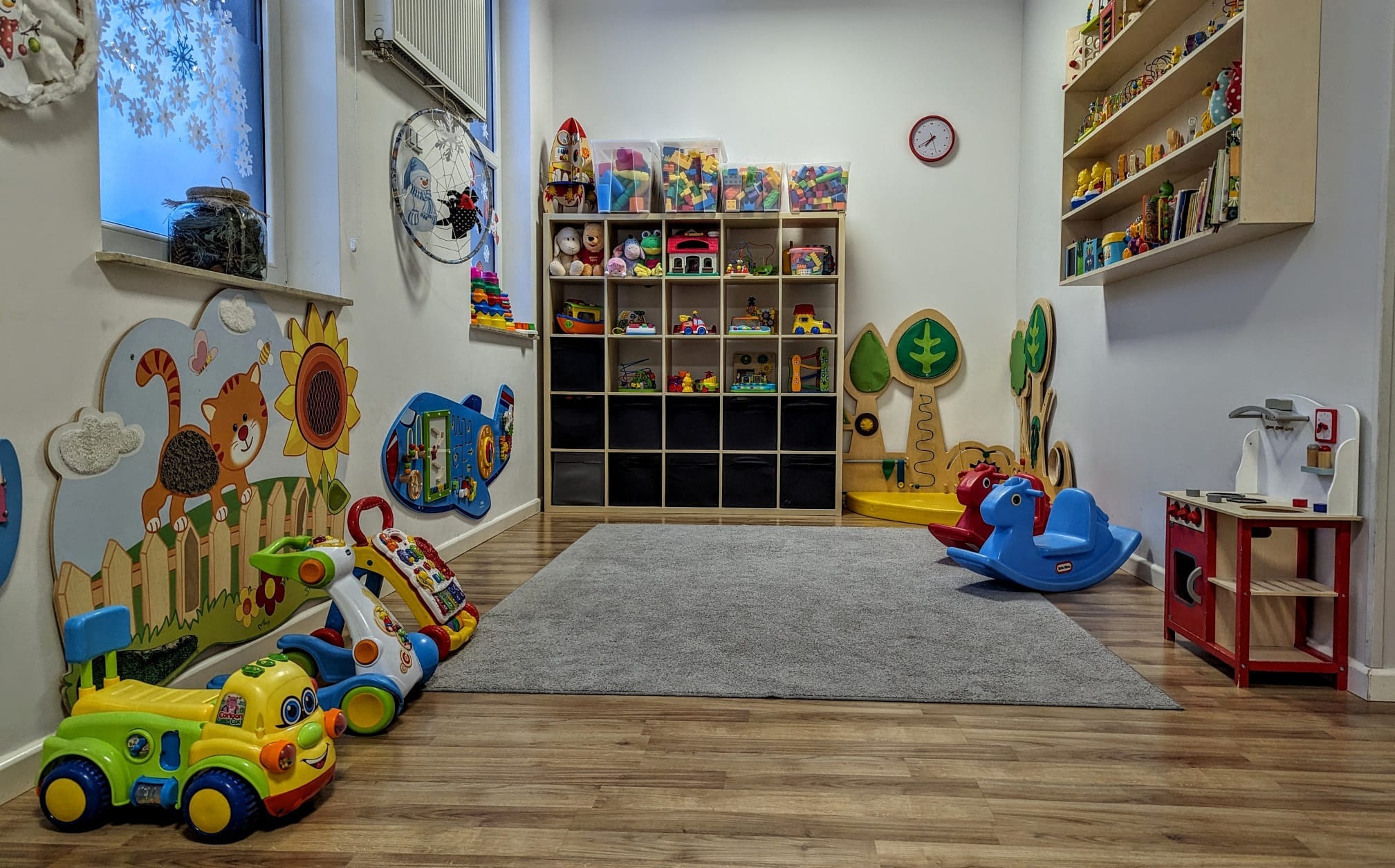 Jasne, przestronne wnętrze sali zabaw z drewnianą podłogą, szarą wykładziną i kolorowymi zabawkami, w tym bujakami w kształcie koników i interaktywnymi pojazdami.