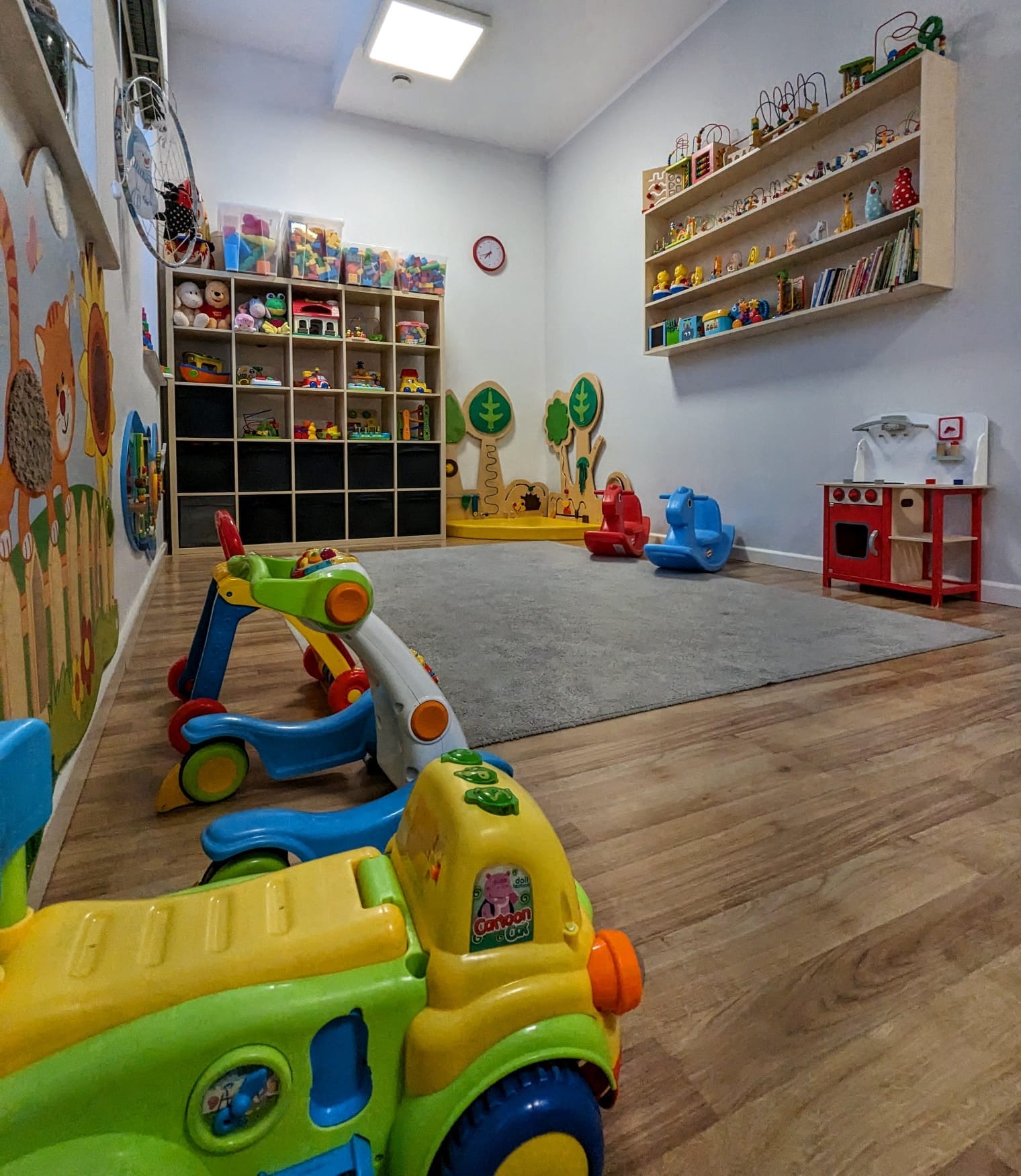 Wnętrze sali zabaw z drewnianą podłogą, szarym dywanem, regałami z zabawkami, drewnianą kuchnią i bujakami w kształcie koników.