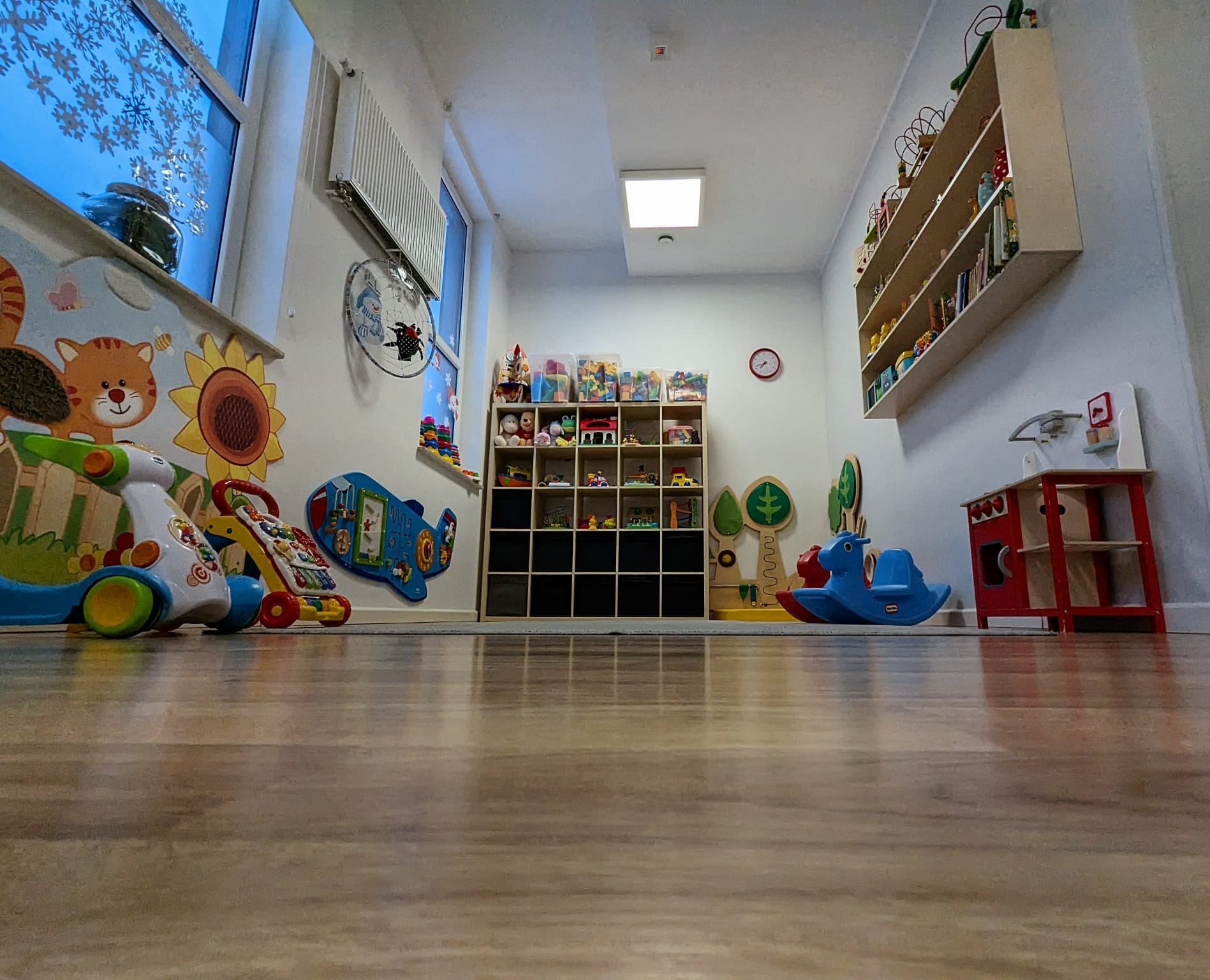 Wnętrze pokoju zabaw z niskiej perspektywy, widoczne zabawki edukacyjne, regał z pojemnikami, malowidła na ścianie i drewniana podłoga.
