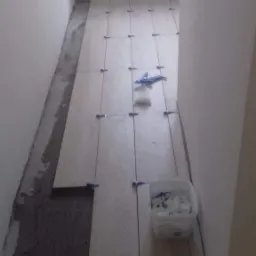 Układanie jasnych płytek podłogowych w wąskim korytarzu, z widocznym klejem, krzyżykami dystansowymi i pojemnikiem na pozostałości po glazurze.