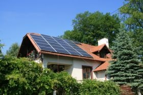 Dom jednorodzinny z panelami słonecznymi na dachu, otoczony zielenią, w słoneczny dzień.