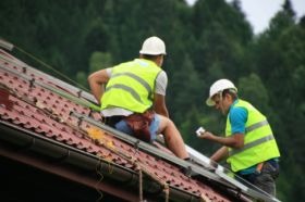 Dwóch monterów w kaskach i kamizelkach odblaskowych instaluje panele na dachu pokrytym czerwoną dachówką, z widocznym orynnowaniem i zielonym tłem drzew.