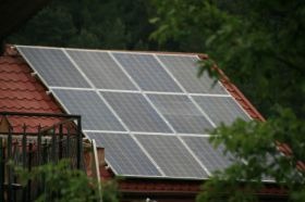 Panele słoneczne na skośnym dachu pokrytym czerwoną dachówką, widoczne zza zieleni drzew.