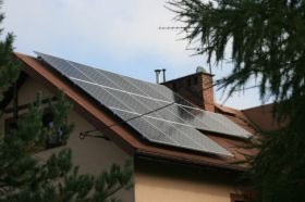 Panele słoneczne na dachu domu jednorodzinnego, widoczne kominy i drzewa w tle.