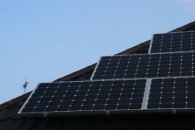 Panele słoneczne zamontowane na ciemnym, spadzistym dachu, widoczne z dołu pod kątem, z błękitnym niebem w tle i fragmentem wiatromierza na szczycie dachu.