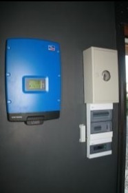 Niebieski inwerter fotowoltaiczny zamontowany na szarej ścianie obok białej skrzynki elektrycznej z bezpiecznikami i licznikiem.