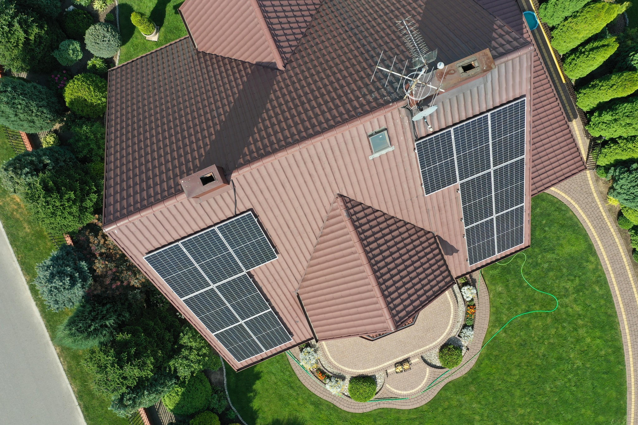 Widok z góry na brązowy dach domu z panelami słonecznymi; dodatkowo widoczna antena satelitarna i zielony trawnik z dekoracyjną ścieżką z kostki.