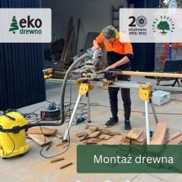 EKODREWNO - Pracownik w odzieży roboczej tnie deskę piłą elektryczną na stojaku. Obok leżą docięte elementy i żółty odkurzacz przemysłowy. W tle widoczne logo firmy.