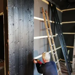 Montaż paneli ściennych z drewna opalanego metodą Yakisugi. Pracownik przykręca czarne deski do ściany za pomocą wkrętarki, obok stoi drabina i leżą dodatkowe deski.