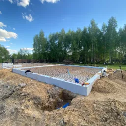 Wylany fundament z widocznym zbrojeniem, przygotowany pod budowę domu. W tle drzewa i błękitne niebo z chmurami.