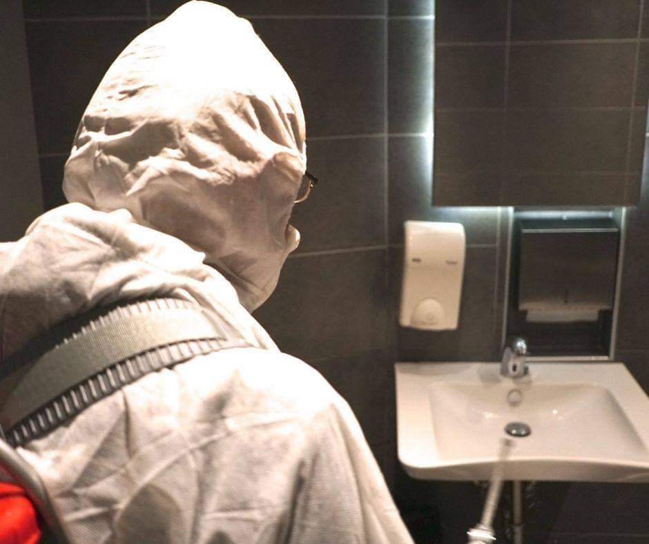 Osoba w kombinezonie ochronnym i okularach, z widocznym fragmentem czerwonej torby, stoi w łazience z ciemnymi płytkami i białą umywalką, prawdopodobnie przygotowując się do pracy.