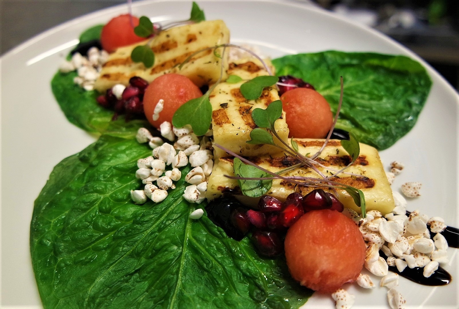 Elegancko podane danie: grillowany ser halloumi z granatem, arbuzowymi kulkami, listkami sałaty i dmuchanym ryżem, polane balsamicznym sosem na białym talerzu.