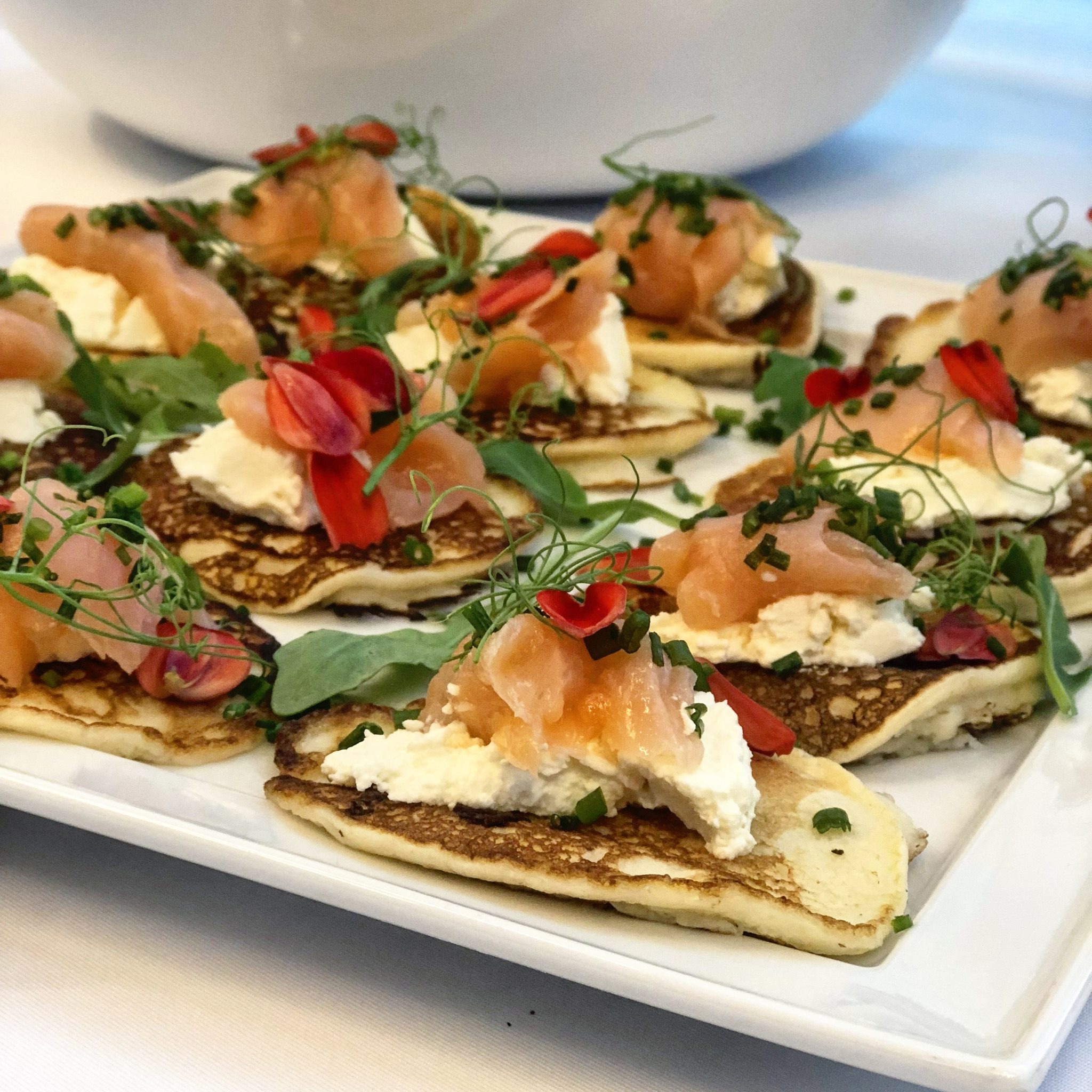 Elegancko podane mini naleśniki z kremowym serem, wędzonym łososiem, rucolą i jadalnymi kwiatami na białym talerzu.