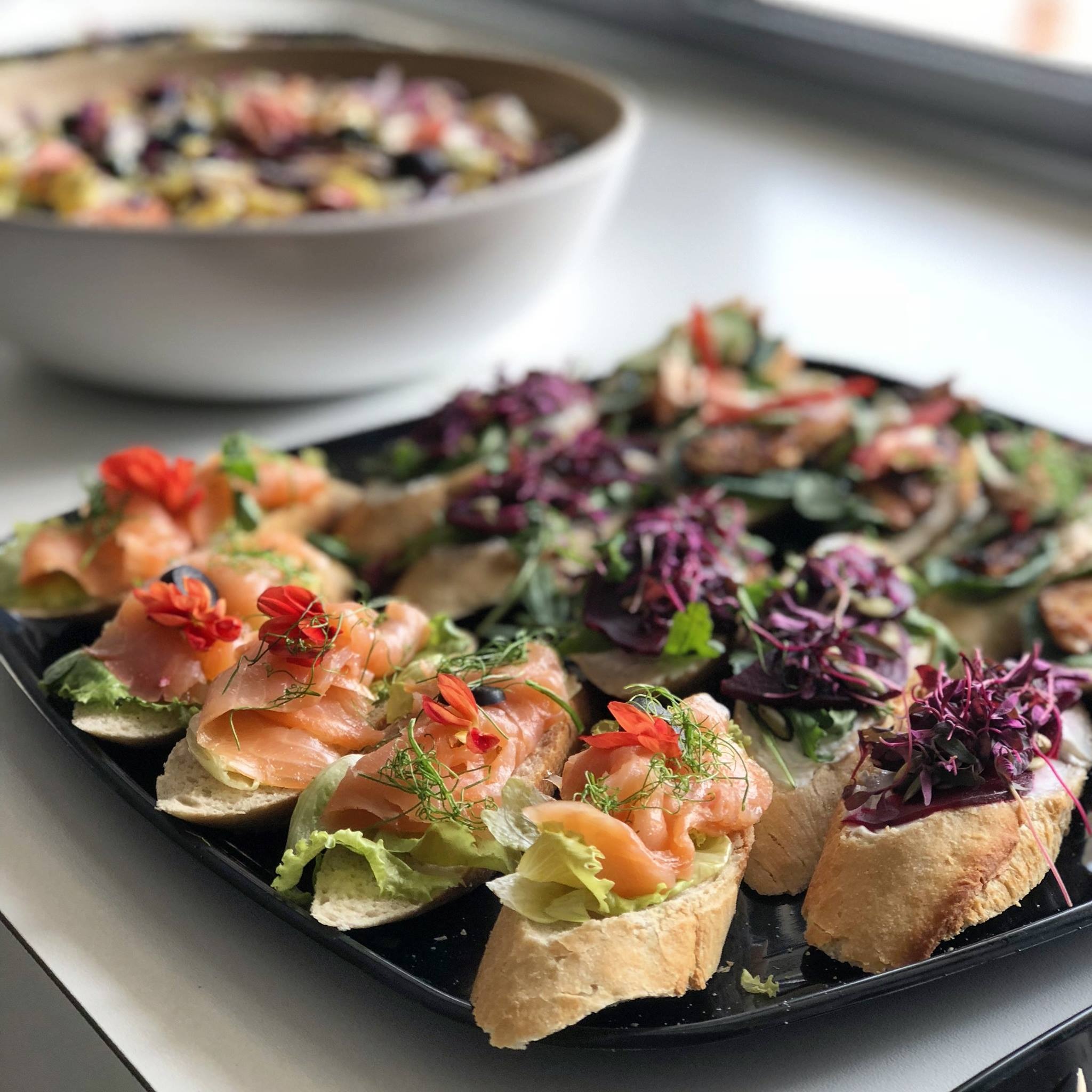 Elegancko podane kanapki na czarnym talerzu: łosoś z sałatą i jadalnymi kwiatami oraz pasta z buraków z fioletowymi kiełkami, w tle miska z sałatką.