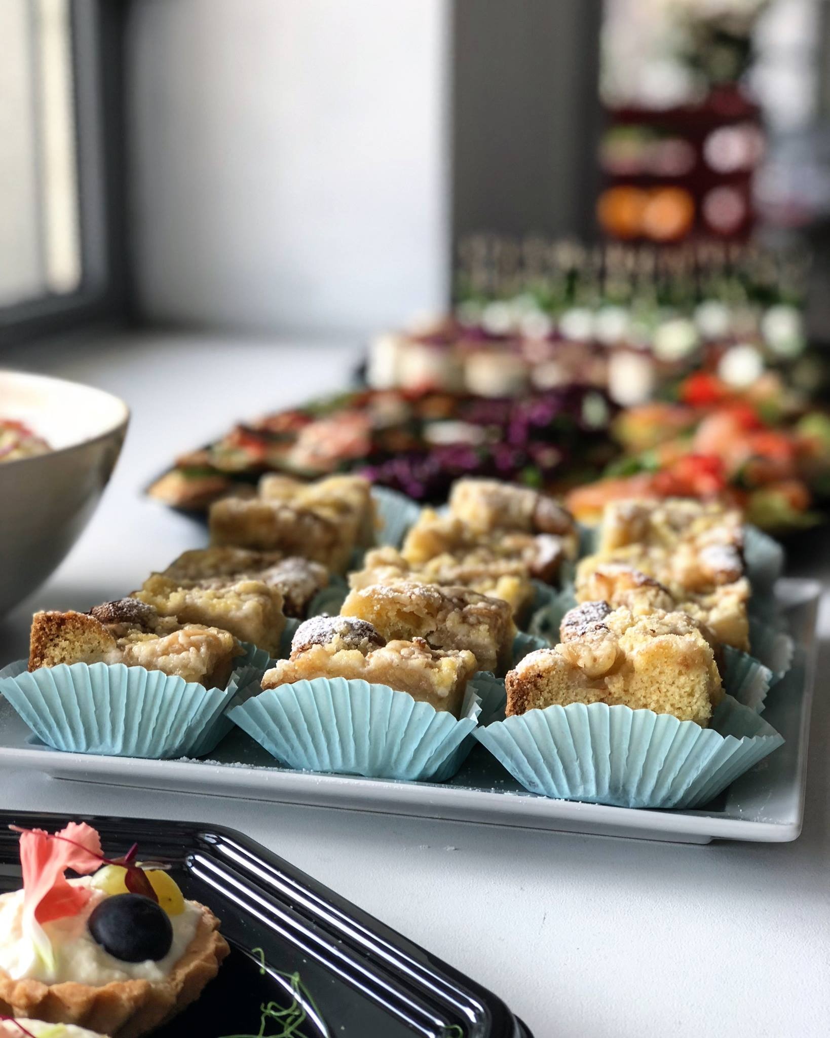 Rząd ciast jabłkowych w błękitnych papilotkach, oprószony cukrem pudrem, na białym talerzu, z innymi przekąskami w tle, w tym tartaletki z owocami i kanapki.