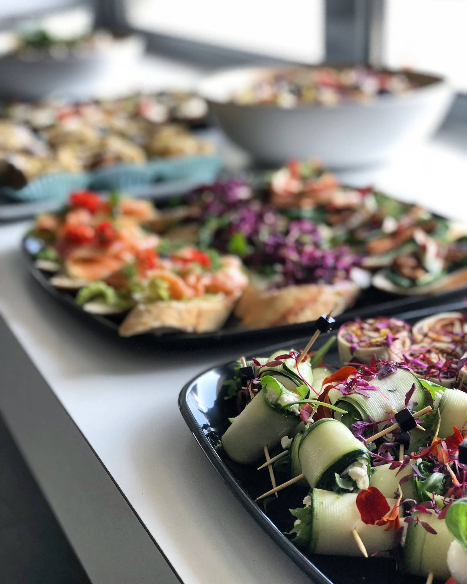 Elegancko podane przekąski cateringowe: rolki z ogórka z białym nadzieniem, mini-kanapeczki z łososiem i sałatki w misach na białym stole.