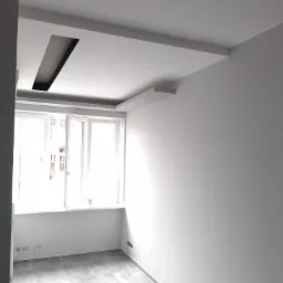 Jasne wnętrze pokoju z szarymi płytkami podłogowymi i białymi ścianami, z oknem w tle i podwieszanym sufitem z ciemnym wnęką na oświetlenie.