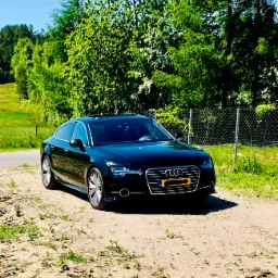 Czarny samochód osobowy marki Audi zaparkowany na piaszczystej drodze polnej, w tle zielone drzewa i krzewy oraz fragment metalowej siatki ogrodzeniowej.