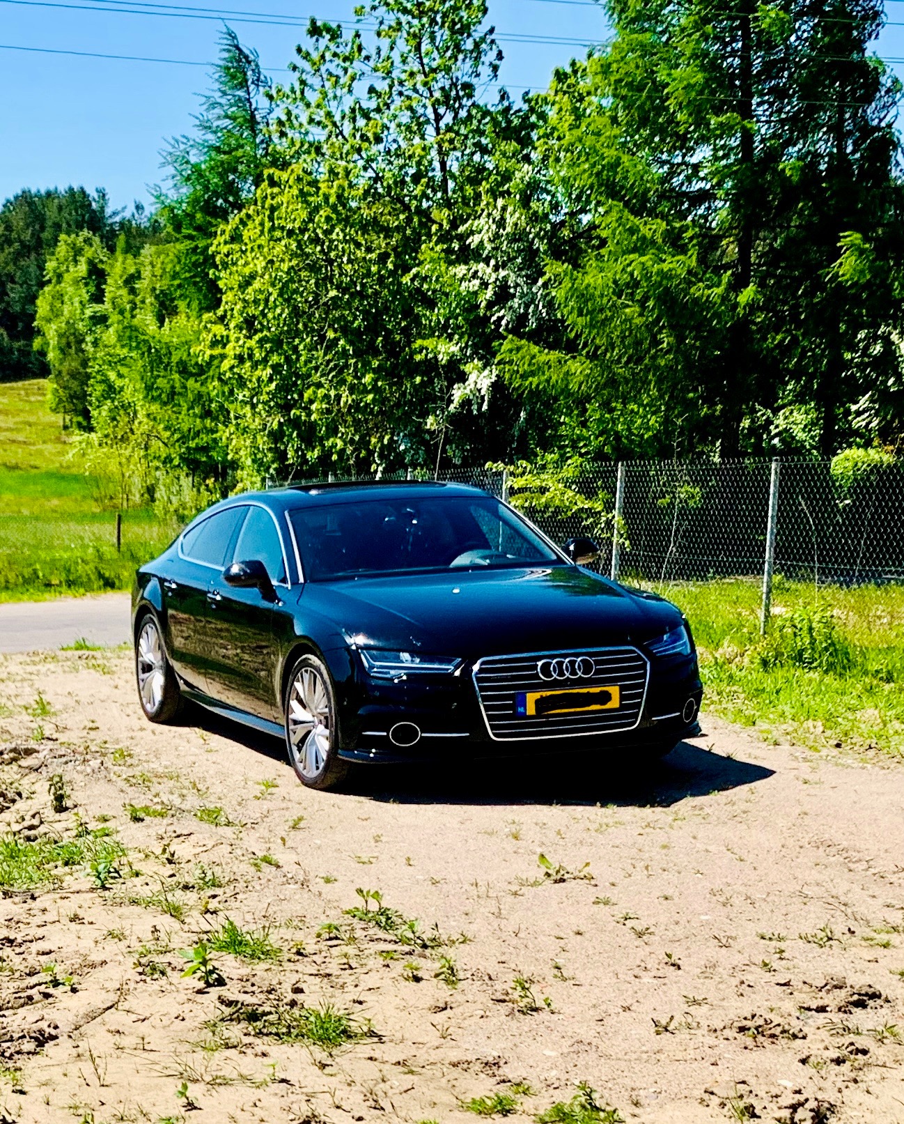 Czarny samochód osobowy marki Audi zaparkowany na piaszczystej drodze polnej, w tle zielone drzewa i krzewy oraz fragment metalowej siatki ogrodzeniowej.