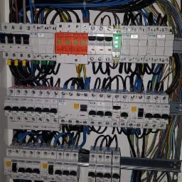 Rozbudowana tablica elektryczna z licznymi bezpiecznikami, przekaźnikami i okablowaniem w kolorach czarnym, niebieskim, żółto-zielonym i brązowym. Widoczne elementy marki Eaton i Hager, z zieloną...