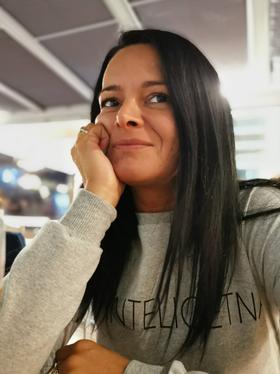 Kobieta z czarnymi włosami, ubrana w szarą bluzę, zamyślona spogląda w górę, opierając brodę na dłoni z pierścionkiem.