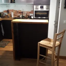 Nowoczesna kuchnia z czarnymi szafkami, drewnianym blatem wyspy i ceglaną ścianą nad blatem, oświetlona trzema wiszącymi lampami z żarówkami Edisona, z drewnianym krzesłem barowym z siedziskiem...