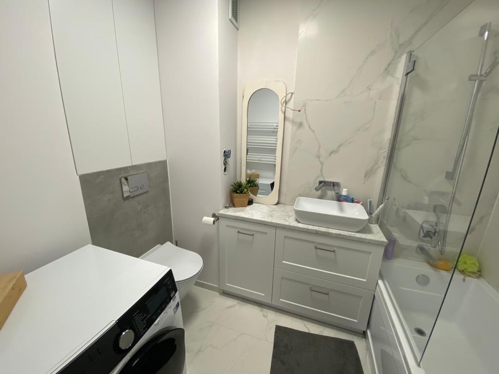 Nowoczesna łazienka z marmurowymi płytkami, białą umywalką nablatową, szafką z szufladami, lustrem w ozdobnej ramie, kabiną prysznicową i pralką.