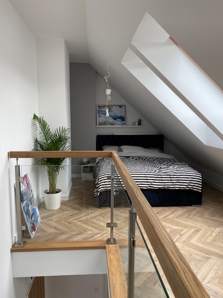 Sypialnia na poddaszu z łóżkiem w czarno-białe pasy, parkietem w jodełkę i szklaną balustradą na antresoli, oświetlona wiszącą lampą i światłem z okien dachowych.