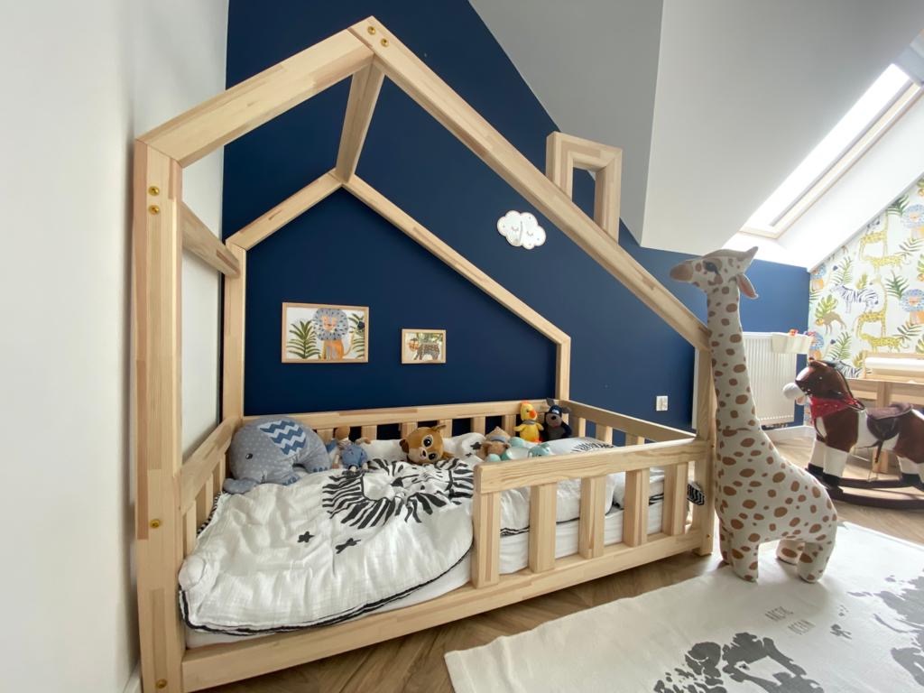 Pokój dziecięcy z drewnianym łóżkiem w kształcie domku, niebieską ścianą i tapetą w zwierzęta, uzupełniony o pluszaki i dekoracje ścienne.