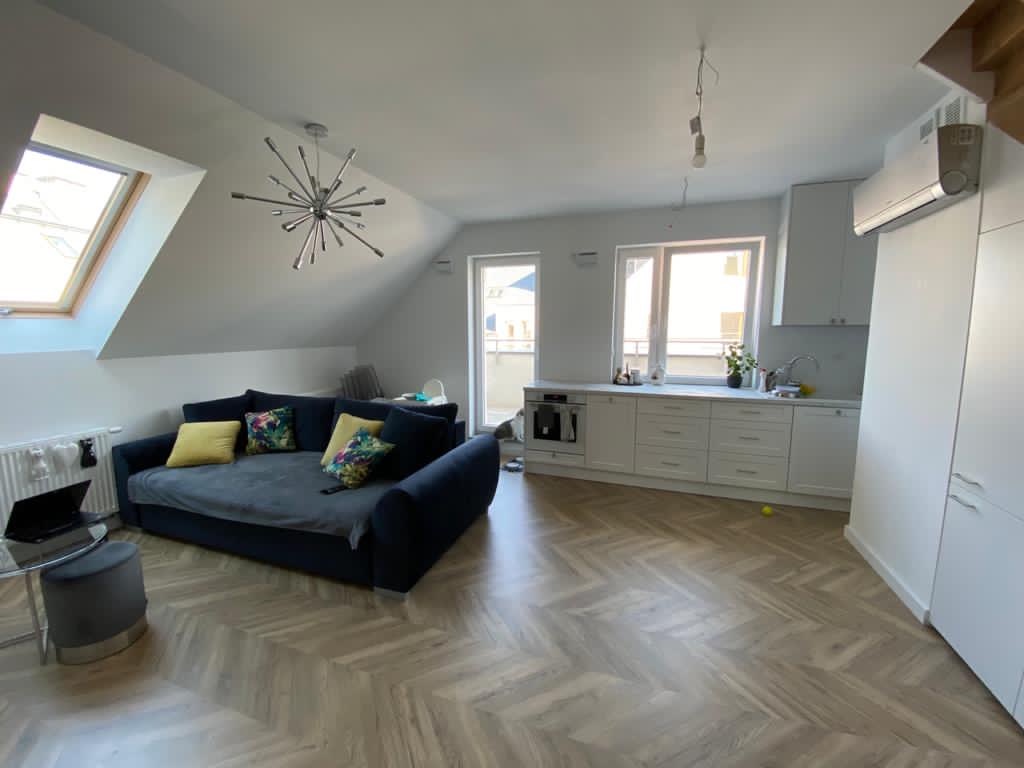 Jasne, przestronne wnętrze poddasza z parkietem w jodełkę, obejmujące aneks kuchenny z białymi szafkami i salon z ciemnoniebieską sofą i dekoracyjnym oświetleniem.