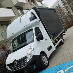 Biały samoch&oacute;d dostawczy Renault Master z czarną plandeką zaparkowany na niebieskim parkingu dla w&oacute;zk&oacute;w sklepowych, na tle nowoczesnego bloku mieszkalnego z balkonami.