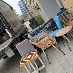 Otwarta paka busa z widocznymi panelami wewnętrznymi, obok wystawione meble ogrodowe i biurowe przygotowane do załadunku na chodniku przed budynkiem mieszkalnym.