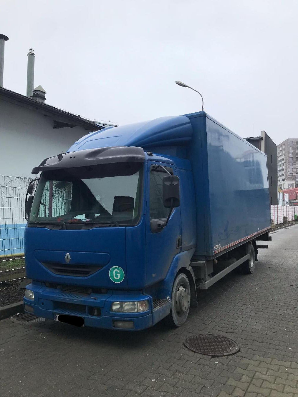 Niebieski samochód ciężarowy Renault z zabudową kontenerową, zaparkowany na szarej kostce brukowej, z widocznym logo 'G' na przedniej masce i lampą uliczną w tle.