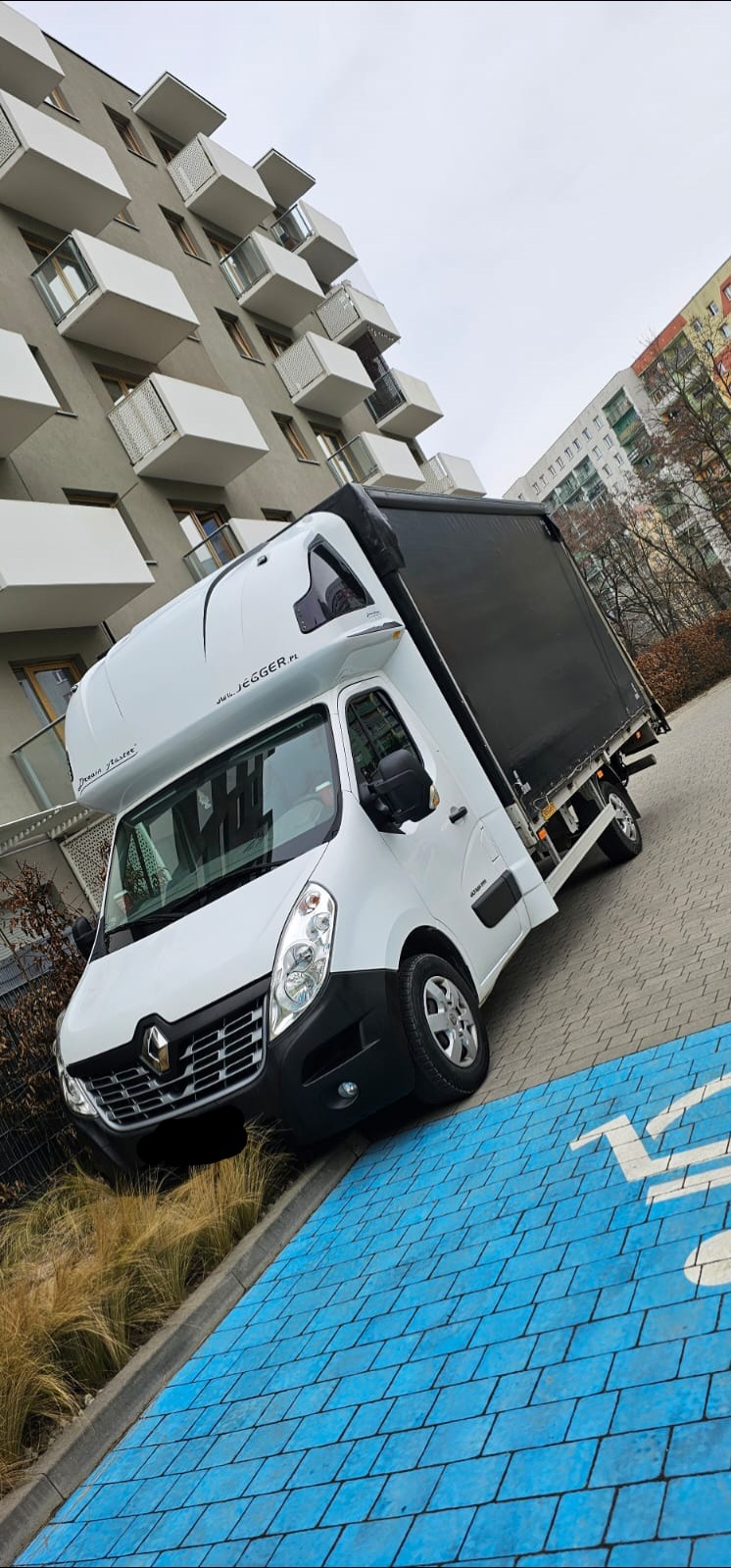Biały samochód dostawczy Renault Master z czarną plandeką zaparkowany na niebieskim parkingu dla wózków sklepowych, na tle nowoczesnego bloku mieszkalnego z balkonami.